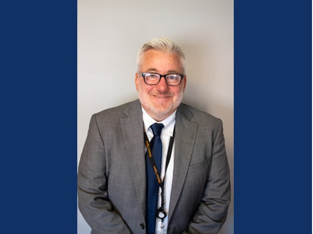 Man with short silver hair and beard wearing glasses, gray suit jacket, blue tie over white collared shirt and a lanyard around his neck.