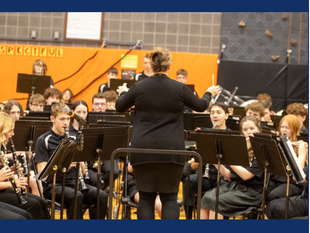 Students sitting on stage playing instruments while person in balck suit conducts