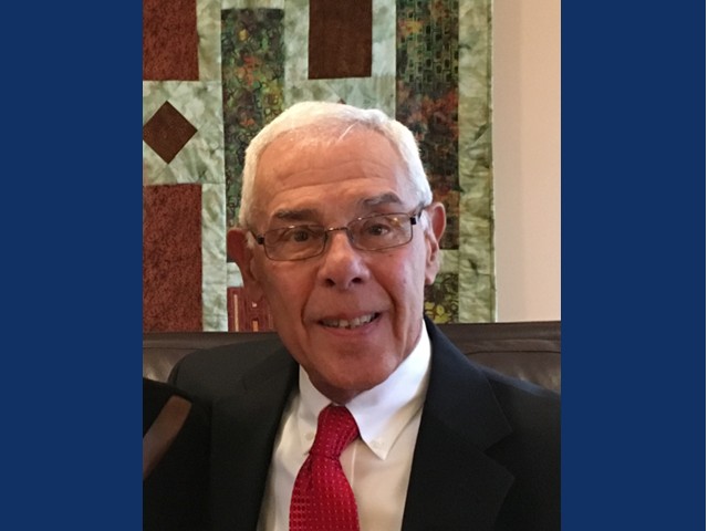 Older man with silver hair, wire glasses in dark suit with red tie