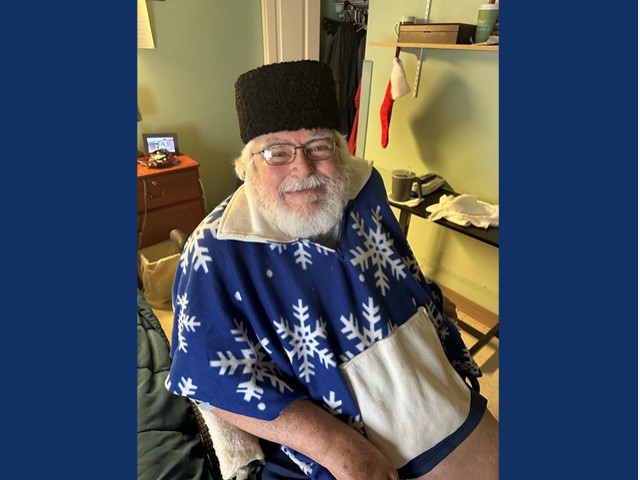 Man with gray beard and mustache with dark rimmed glassed in black straight hat wearing a blue shirt with white snowflakes on it.