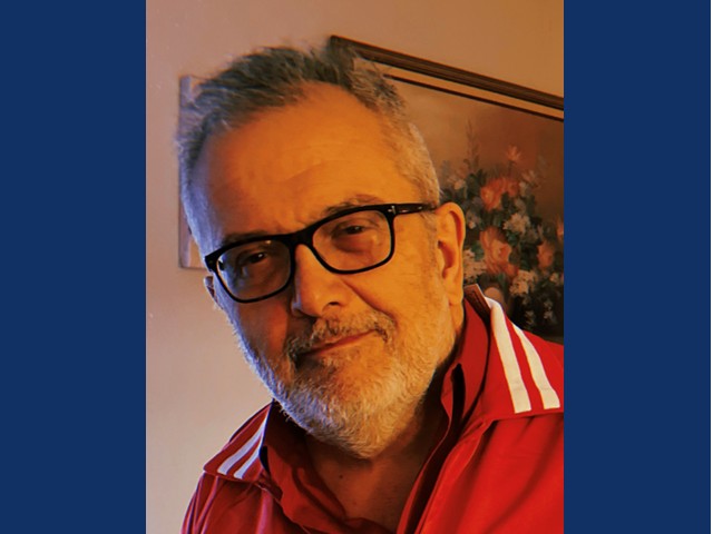 Man with silver hair, dark rimmed glasses smirking in red collared shirt,