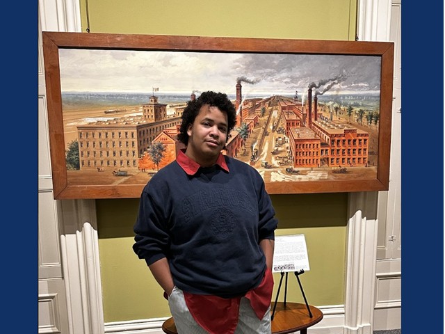 Man in dark blue sweater with red collar in front of large painting of Oswego