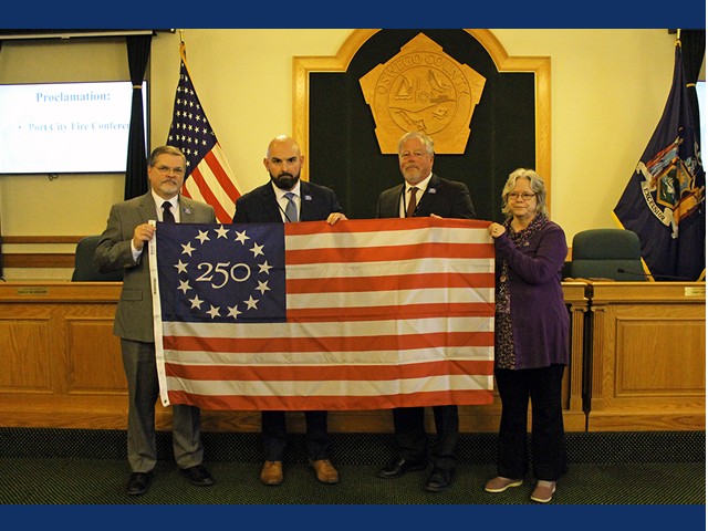 Picture of three men and one woman holding a large flag with 13 stars circling "250" while standing in front of Oswego County emblem.