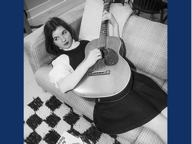 Young dark haired woman in dark dress lying on couch playing acoustic guitar.