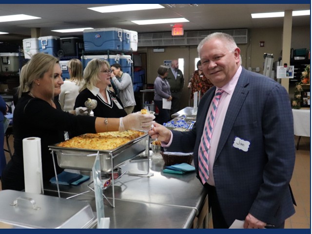 Man with short graying hair in dark suit jacket and pink and blue striped tie receiving a cup from woman with blonde pony tail and black shirt reaching over a silver waarming tray filled with macaroni and cheese.