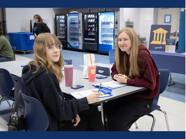 Two blonde girls with long hair and hoodies sitting at a cafeteria table with drinks and paperwork