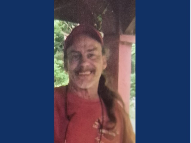 Man withlong brown hair, mustache smiling in cap.