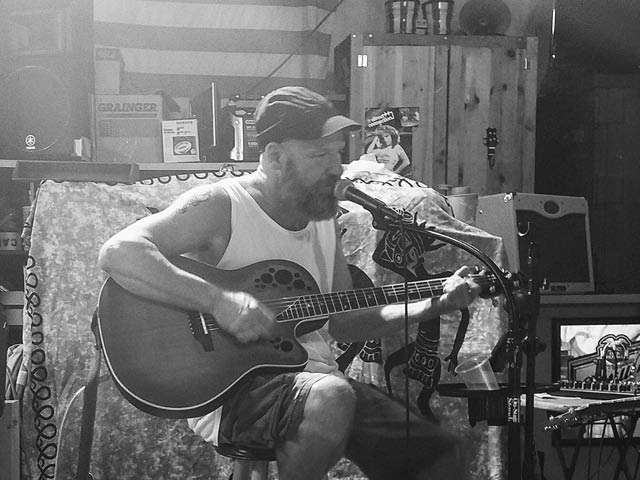 Rich Hart, a white man wearing a baseball hat, tank top and shorts, playing guitar and singing from a seated position