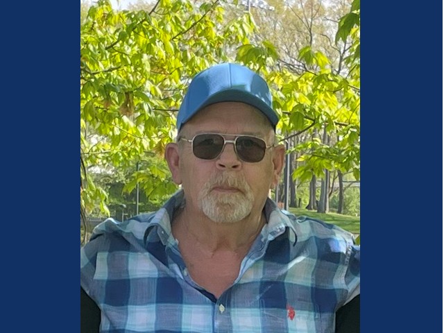 Man with gray short beard and mustache wearing blue and white flannel shirt and blue baseball cap in front of green trees on a sunny day.