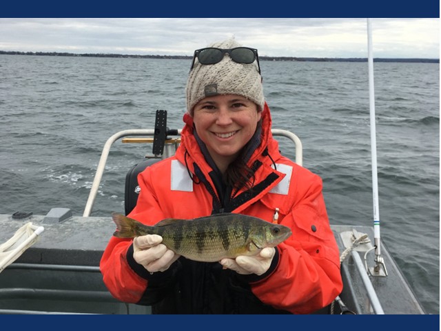 Woman in boat with heavy orange coat, grayish-brown knit cap with sunglasses on her head holding a fresh caught yellow perch in her hands.