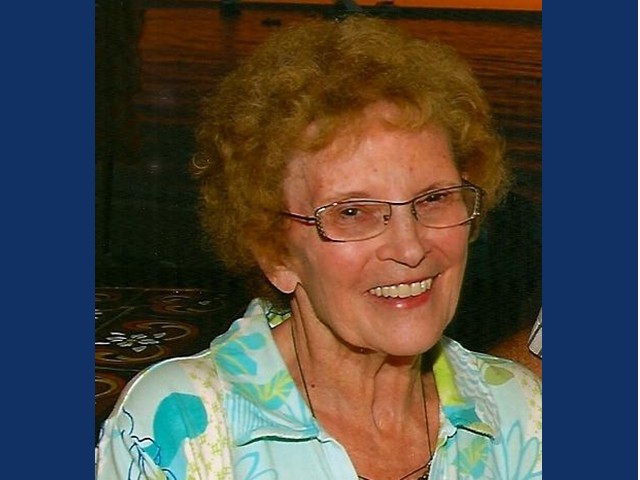 smiling woman with short curly brownish-red hair with glasses and blue print shirt.