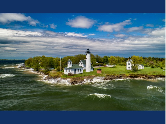 a lighthouse on edge of water with green grass surrounding it, waves coming to shore line. and fluffy white clouds splatter a blue sky
