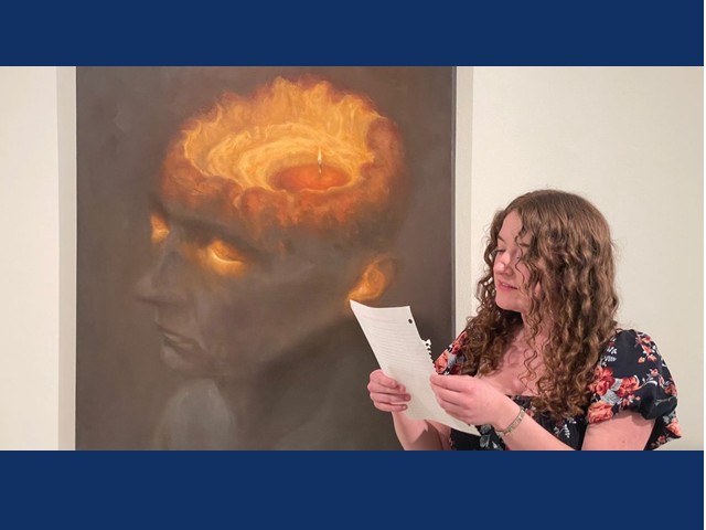 Girl with long curly light brown hair in dark floral shortsleeve reading from white notebook paper in fromt of painting of dark head of person with open top and swirling fire within and glowing from eyes and ears.