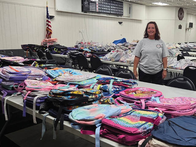 Volunteer standing in front of a table of children's backpacks at last year's Stuff-a-Bus event at the Oswego Elks