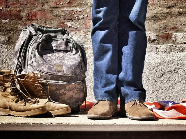 A photo of a person in jeans standing next to a camo backpack, workboots and an American flag