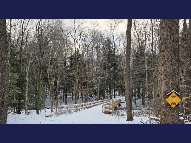 Winona Forest trail with a sign noting a bridge ahead posted to a tree; the wooden walking bridge and trail are both covered in a light dusting of snow